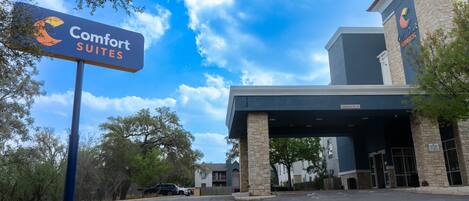Entrada de um hotel Comfort Suites, com céu azul e árvores ao fundo.