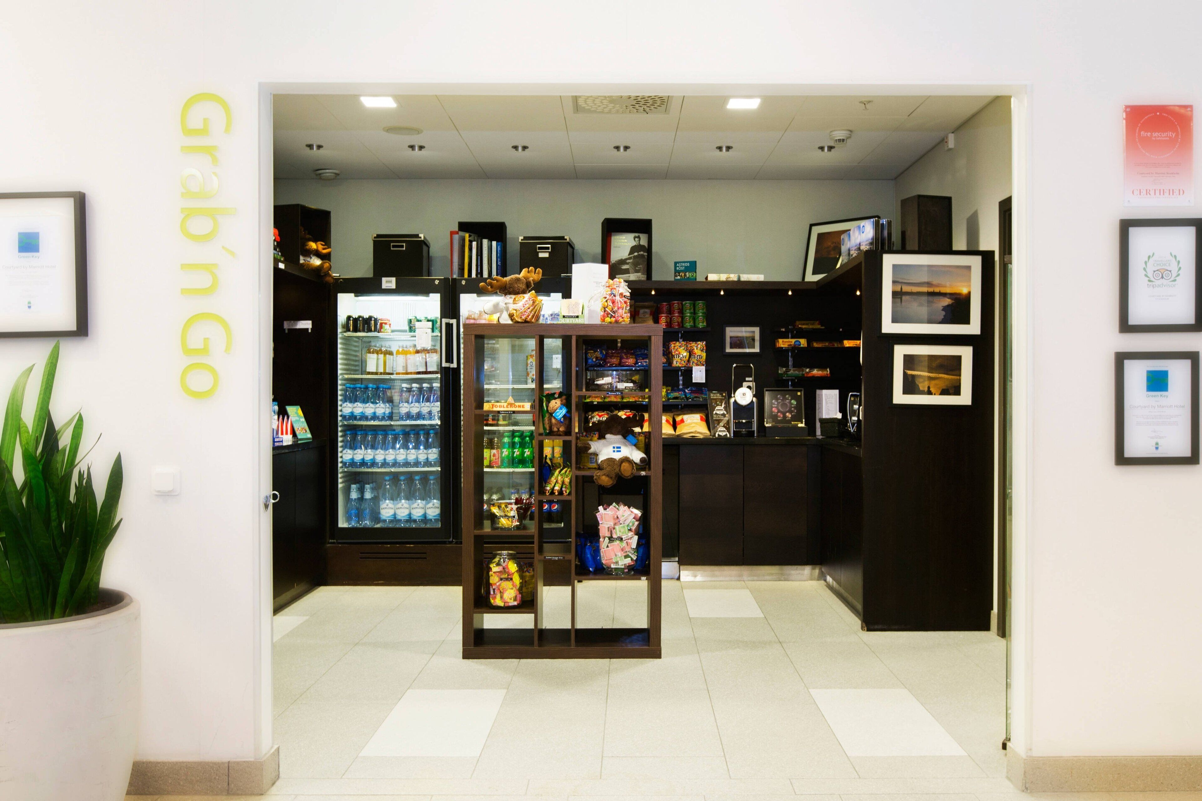 Interior de una tienda con un letrero que dice 'Grab n Go', estantes llenos de bocadillos, un refrigerador con bebidas y fotografías enmarcadas en la pared.