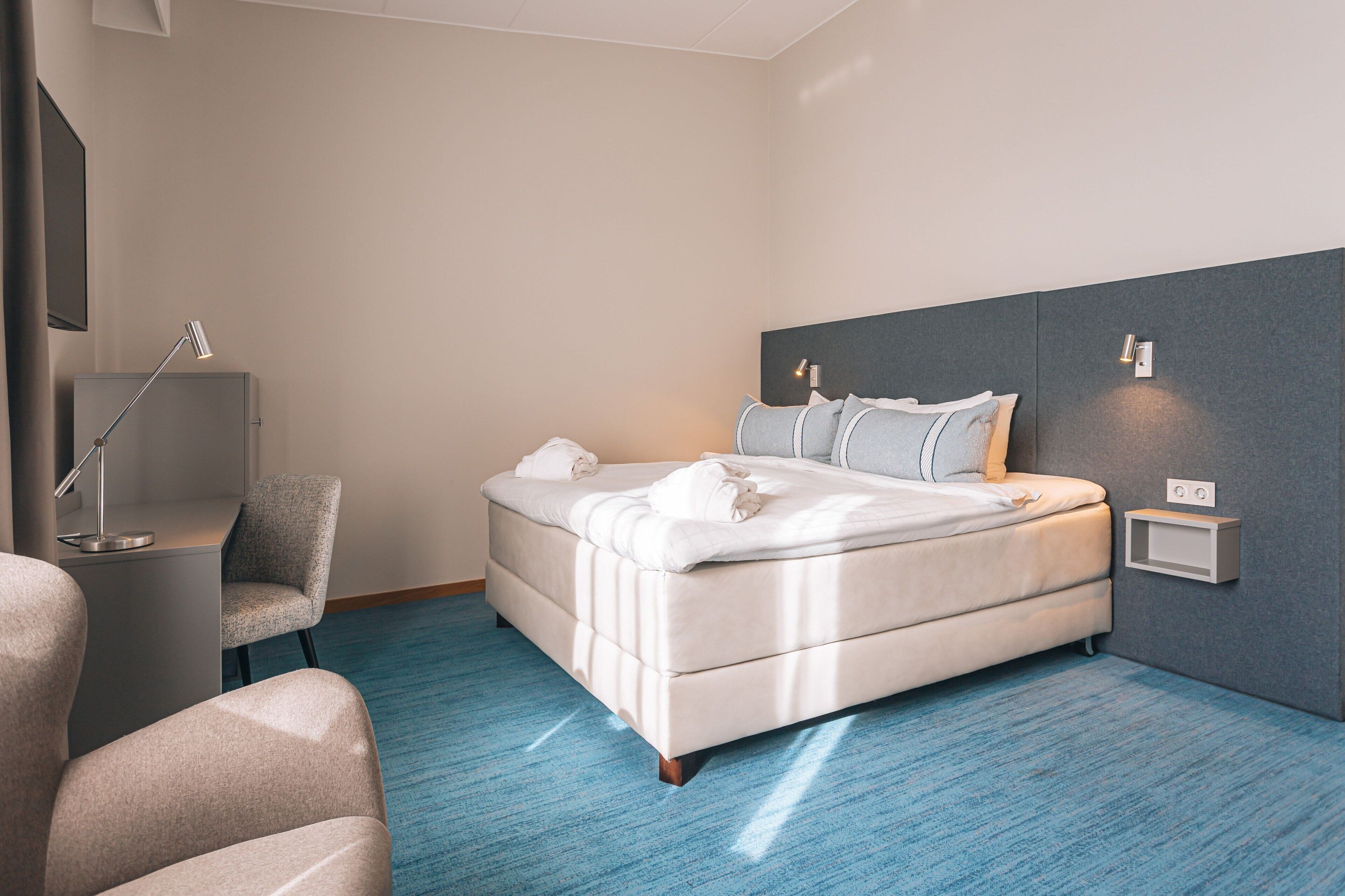 A modern hotel room with a large bed, a desk, a chair, and a wall-mounted light.