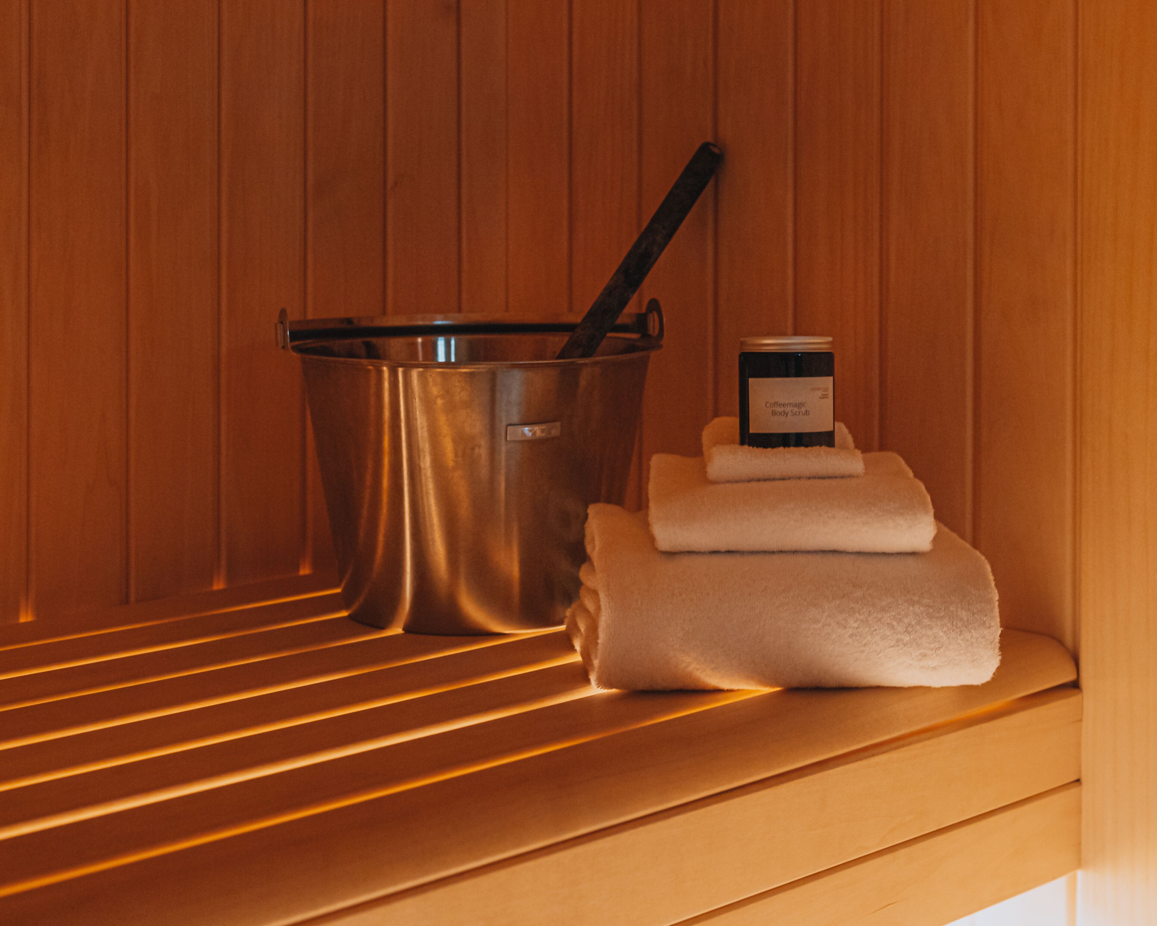 Een sauna met een metalen emmer, een flesje lichaamsscrub en opgevouwen handdoeken op een houten bank.