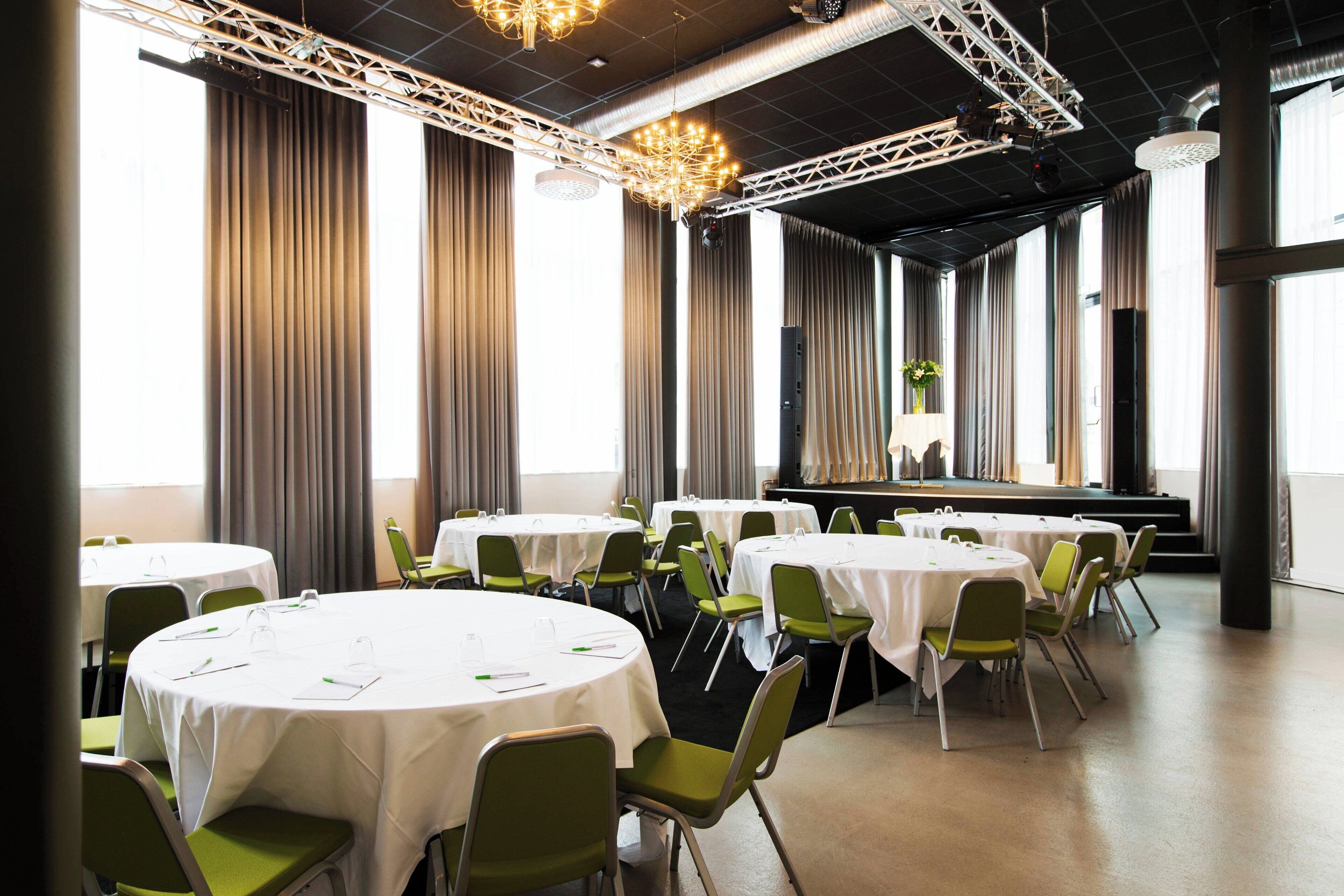 Una sala de conferencias con varias mesas redondas, sillas verdes y un escenario con cortinas.