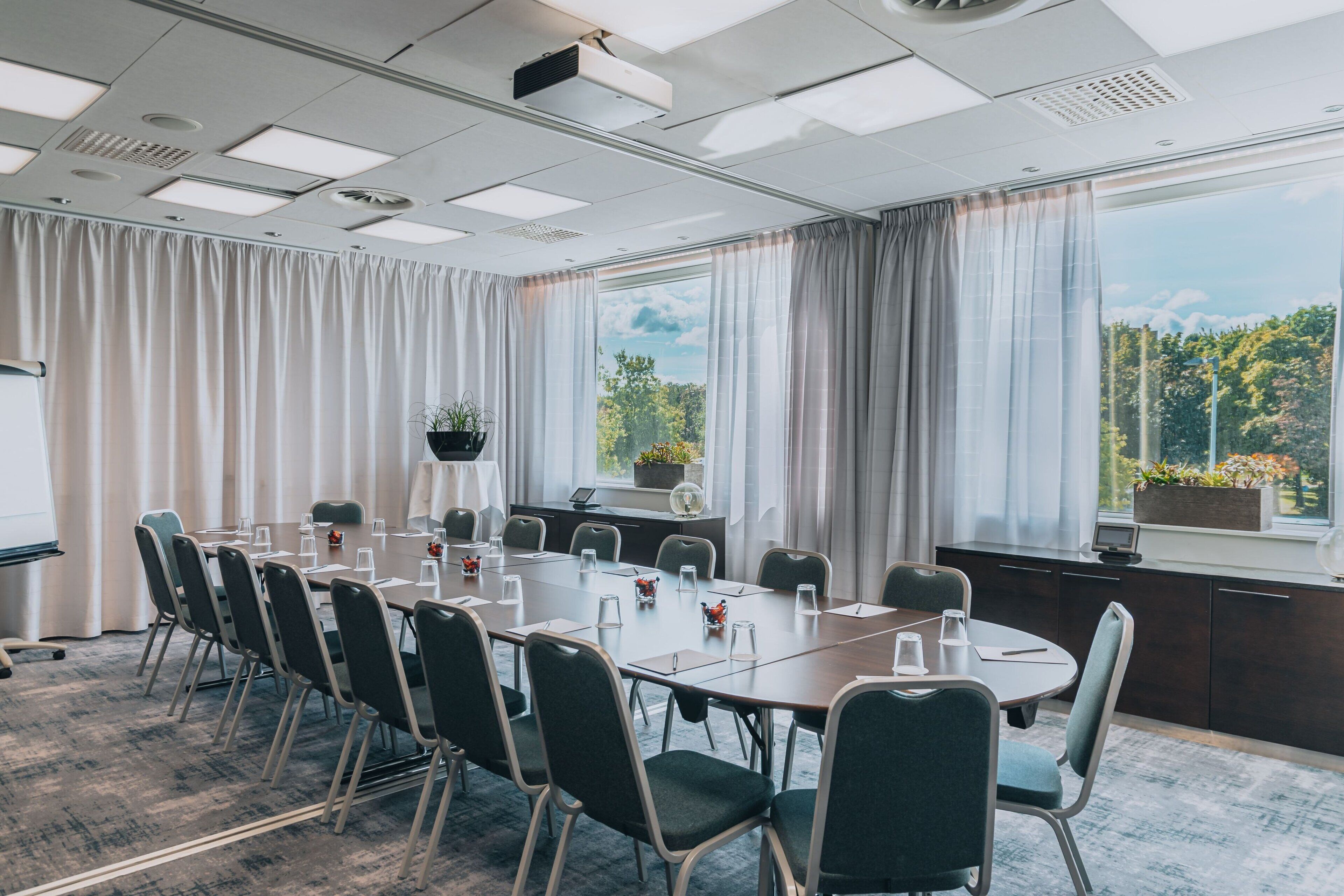 A conference room with a long table, chairs, a whiteboard, and a view of trees through large windows.