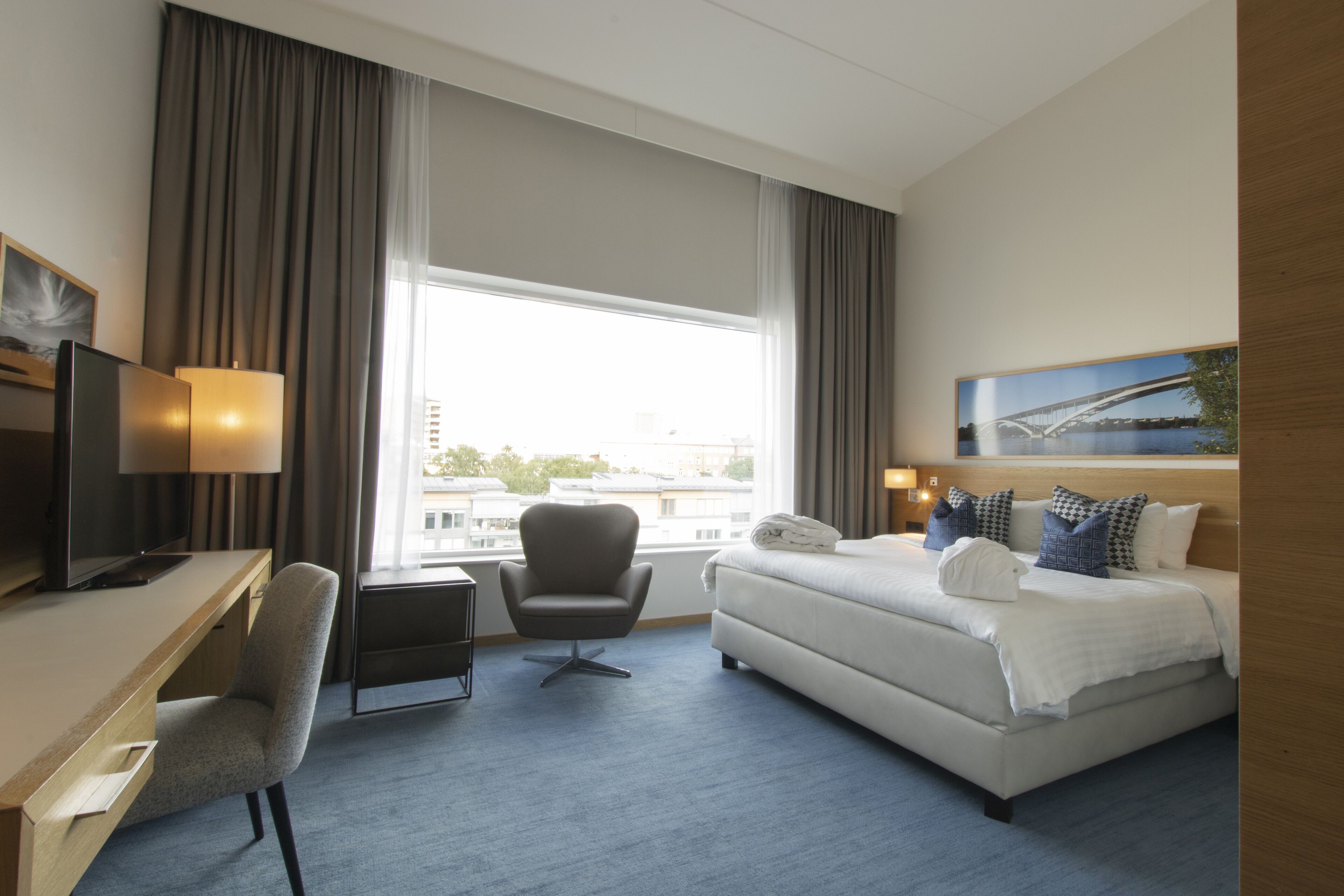 A hotel room with a bed, desk, chair, and large window.