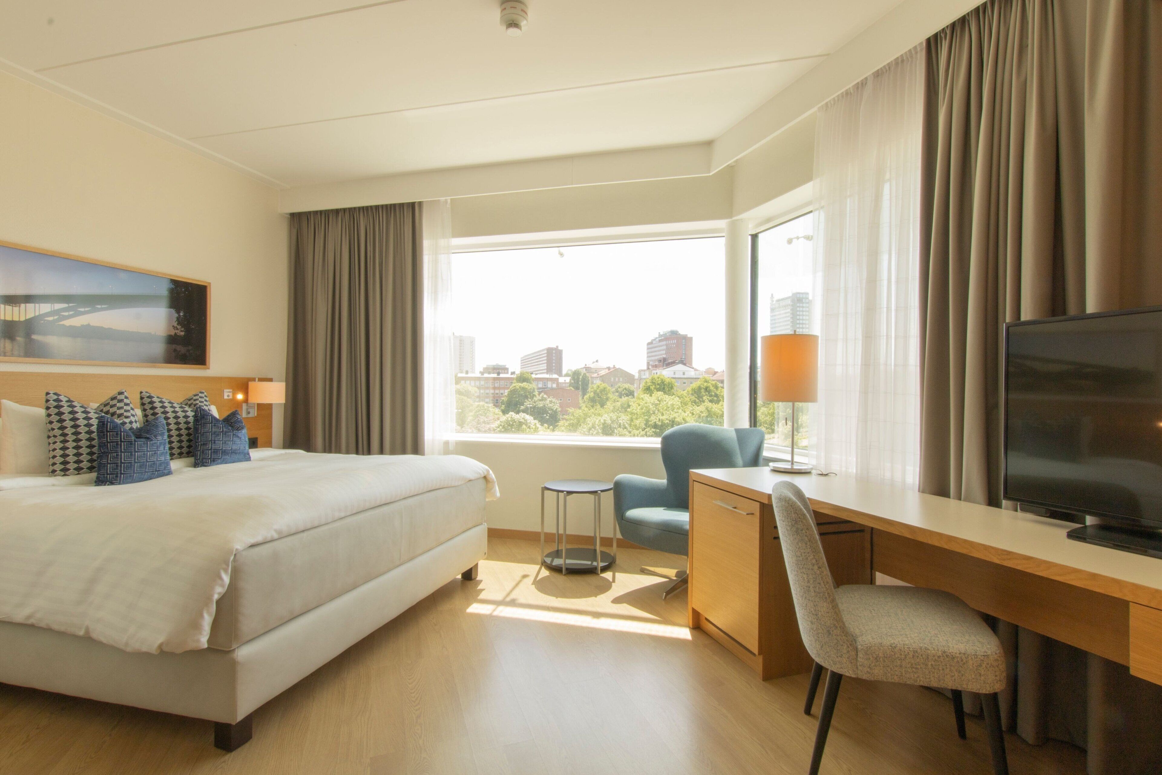 Une chambre d’hôtel comprenant un lit, un bureau, une chaise, un téléviseur et une fenêtre donnant sur des immeubles et des arbres.