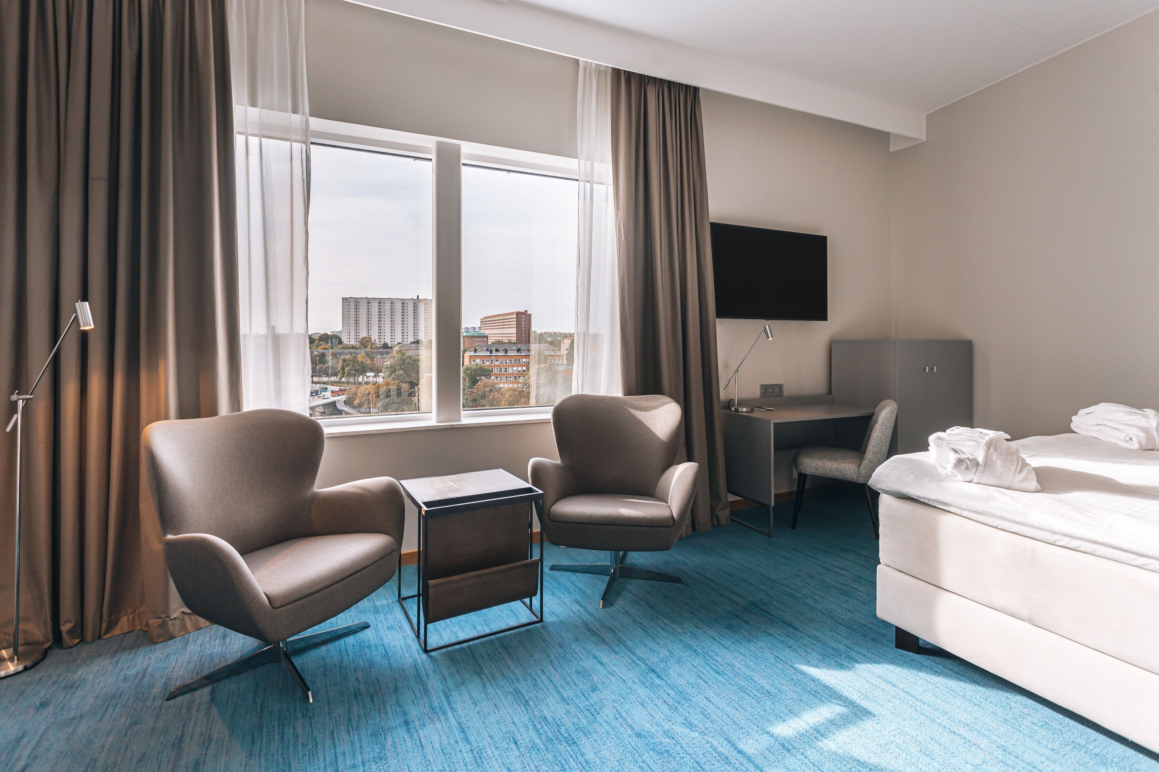 A hotel room with a large window, two armchairs, a desk, a bed, and a flat-screen TV.
