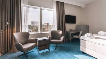 A hotel room with a large window, two armchairs, a desk, a bed, and a flat-screen TV.