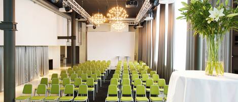 Een vergaderzaal met rijen groene stoelen, een witte tafel met een bloevas en een groot scherm aan de voorzijde.