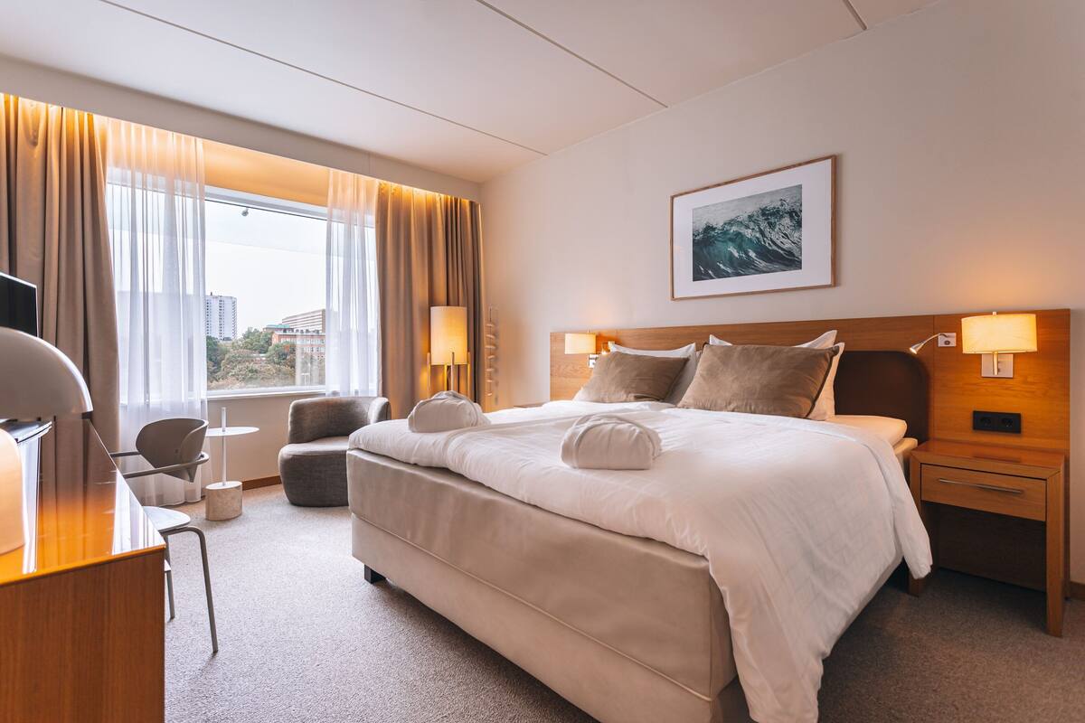 Una habitación de hotel con una cama grande, un escritorio, una silla y un ventanal con cortinas.