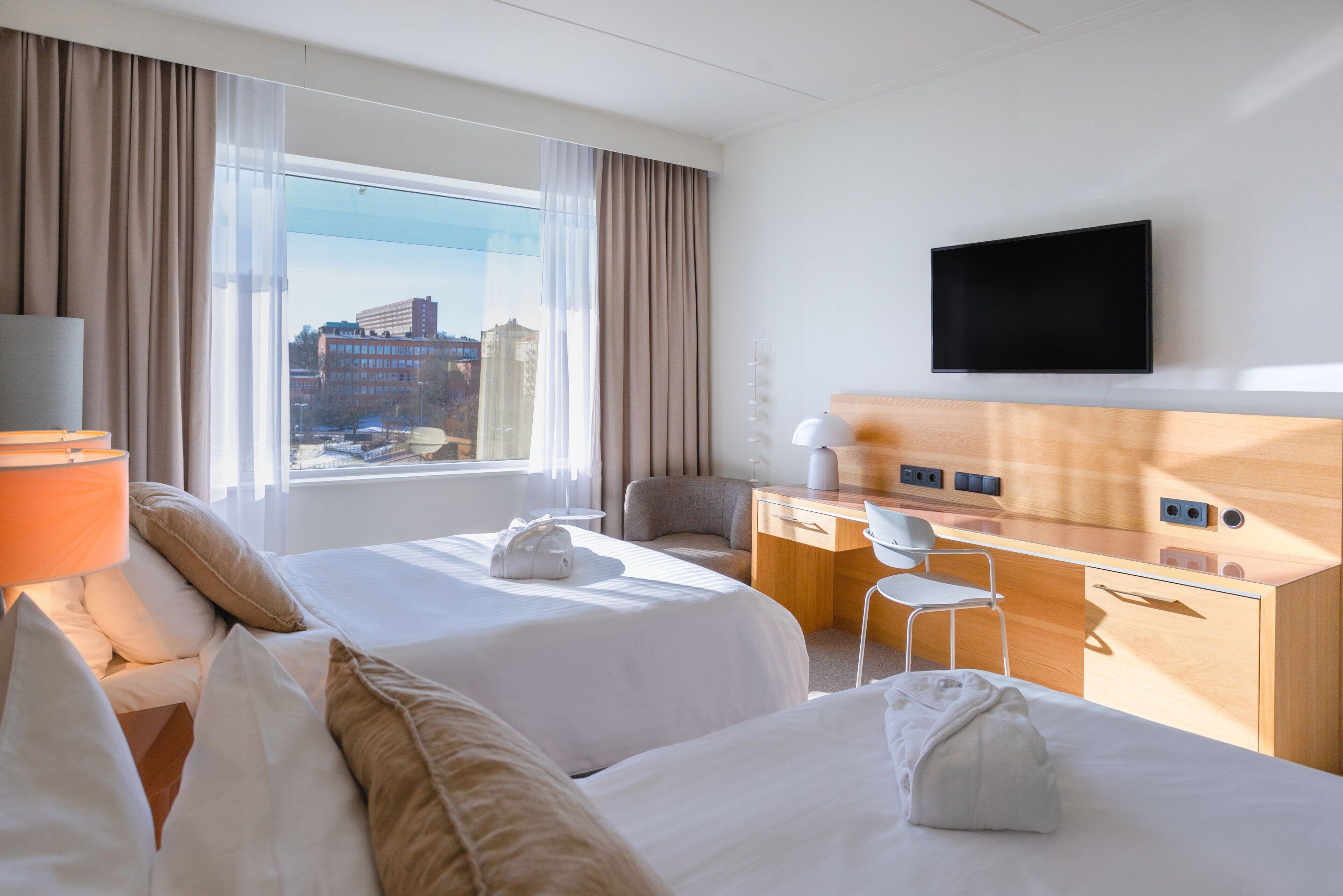 A hotel room with two beds, a desk, a chair, a TV, and a window with a city view.