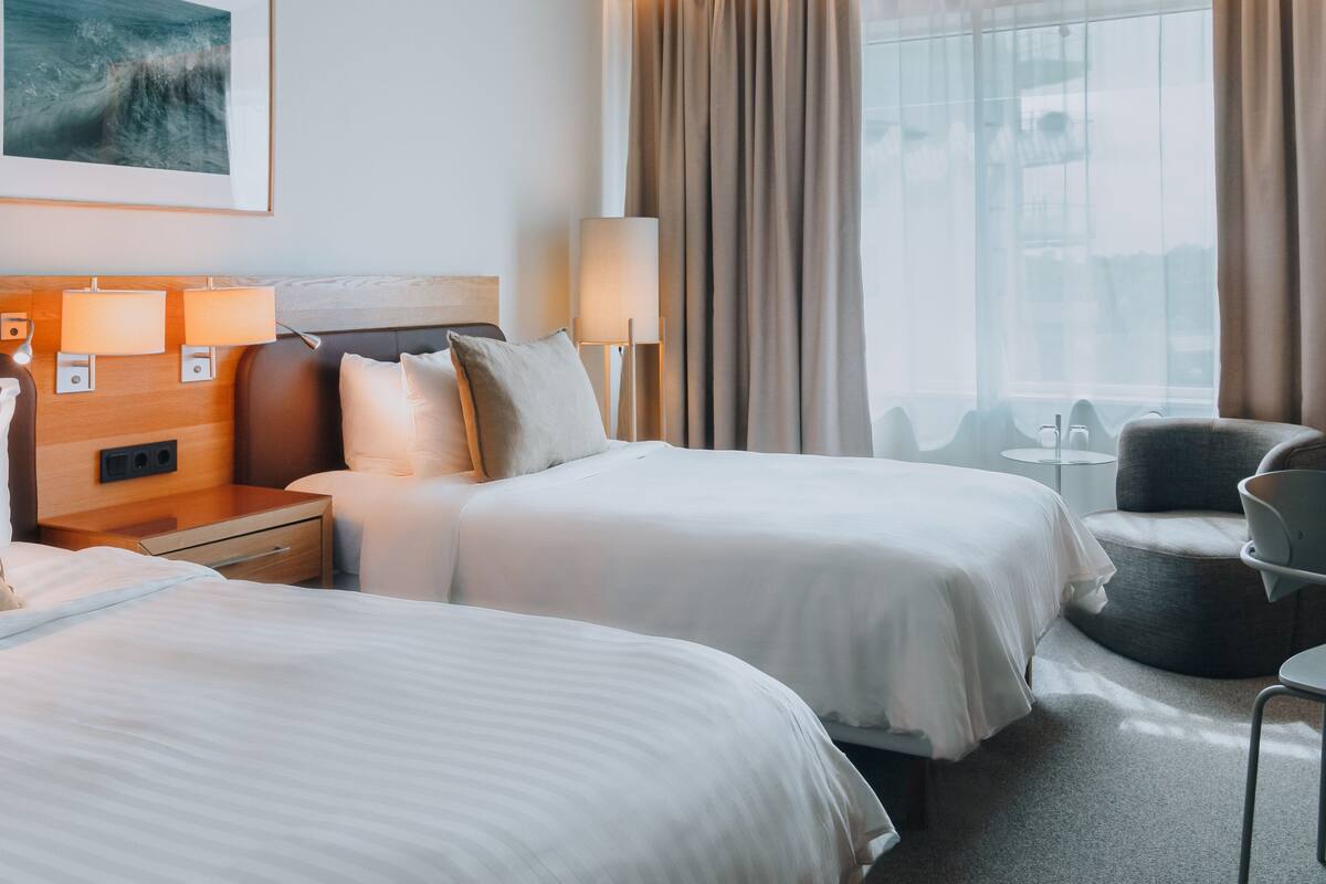 Habitación de hotel con dos camas, un rincón de descanso y un amplio ventanal con cortinas.