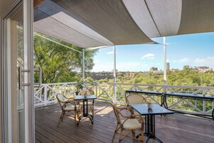 Área externa coberta com mesa e cadeiras, vista para vegetação e edifícios ao longe.