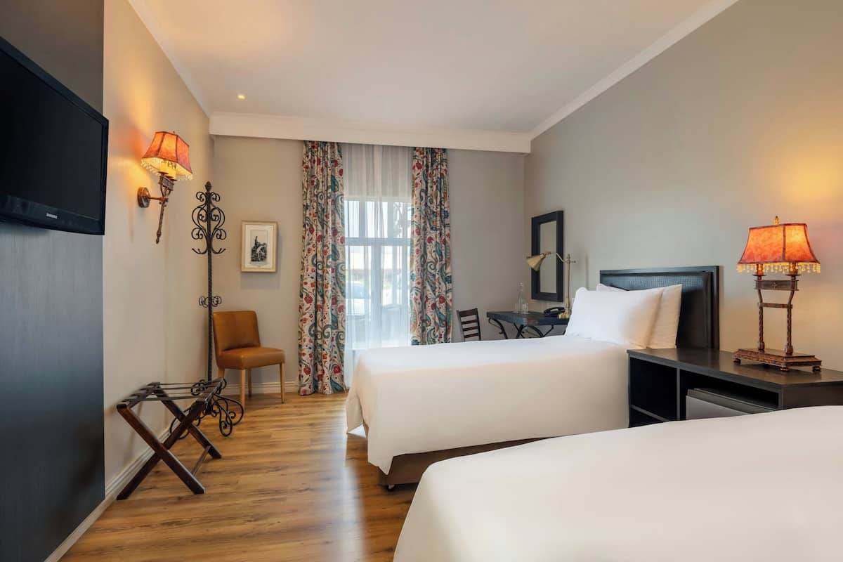 A hotel room with two beds, a desk, a chair, a TV, and two wall-mounted lamps.