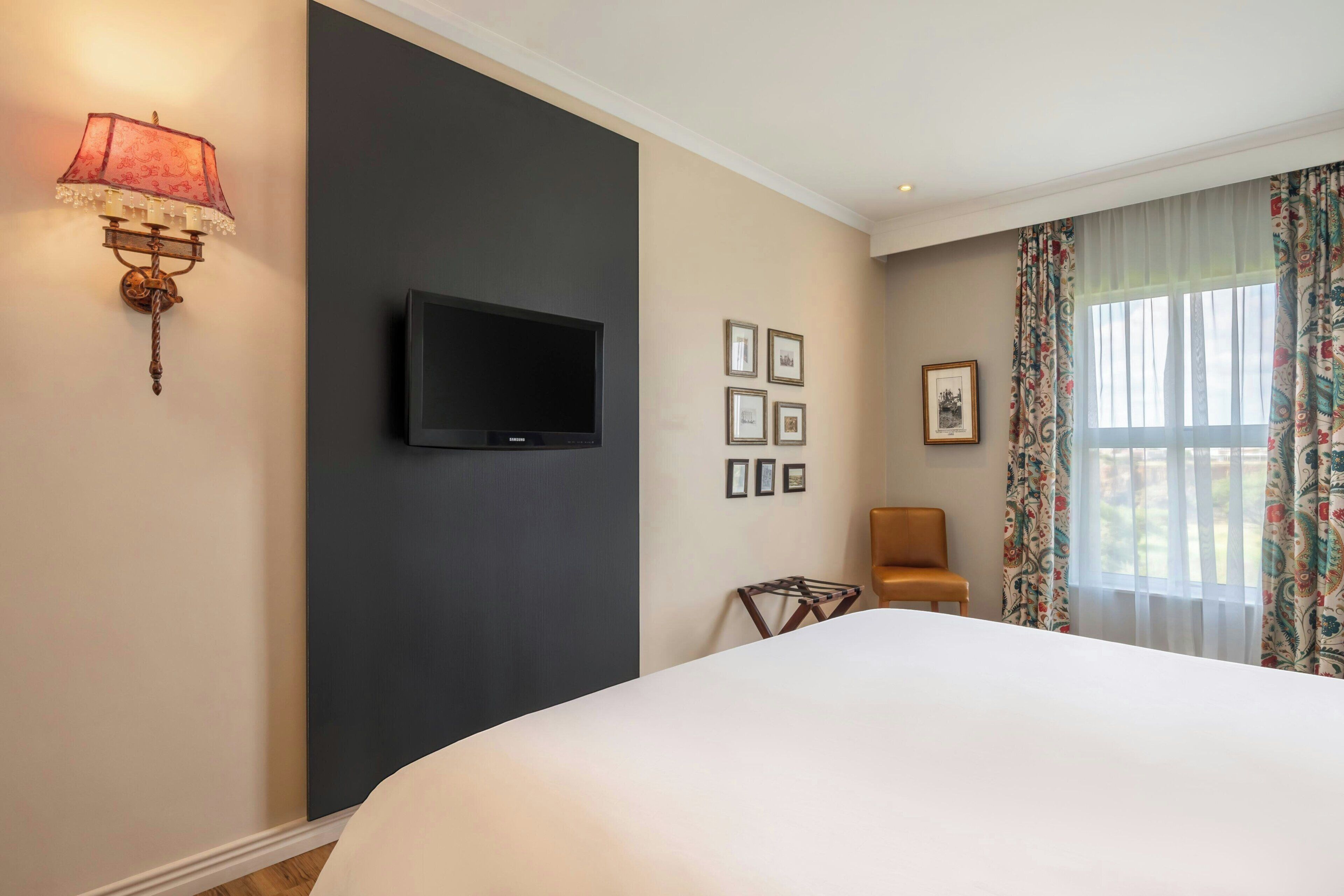A hotel room with a bed, a flat-screen TV, a wall-mounted lamp, a chair, and a window with curtains.