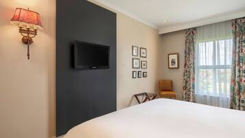 A hotel room with a bed, a flat-screen TV, a wall-mounted lamp, a chair, and a window with curtains.