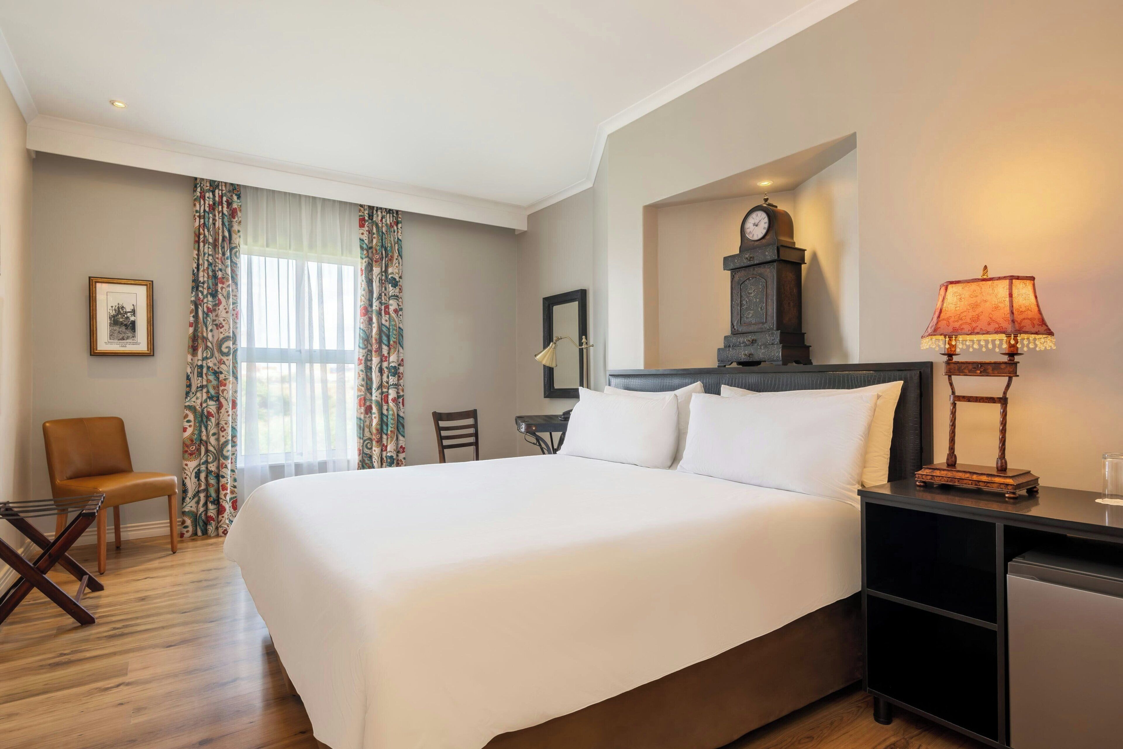 A hotel room with a large bed, a desk with a lamp, a chair, a clock, and a window with curtains.