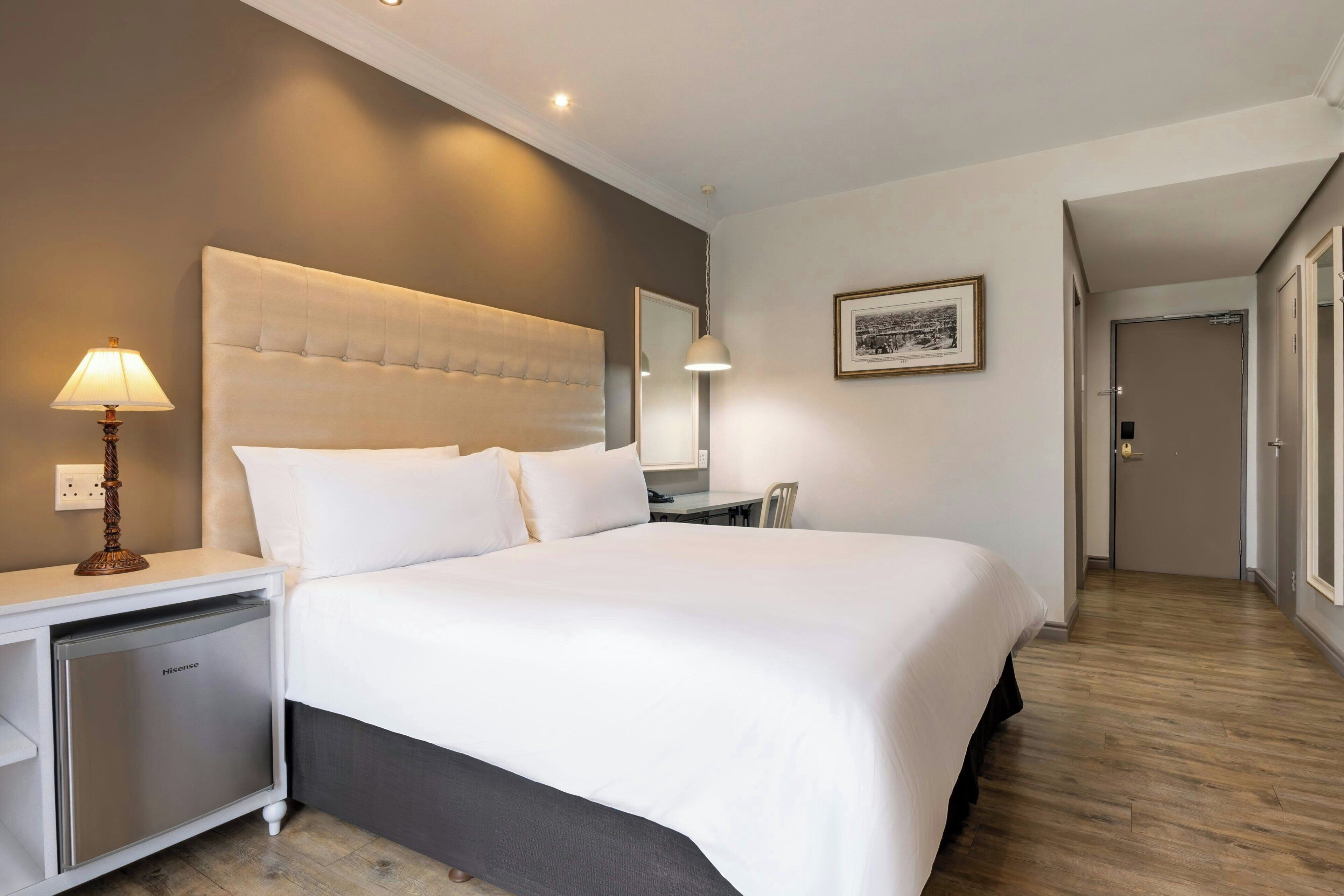 A hotel room with a large bed, bedside table, lamp, and a small refrigerator.