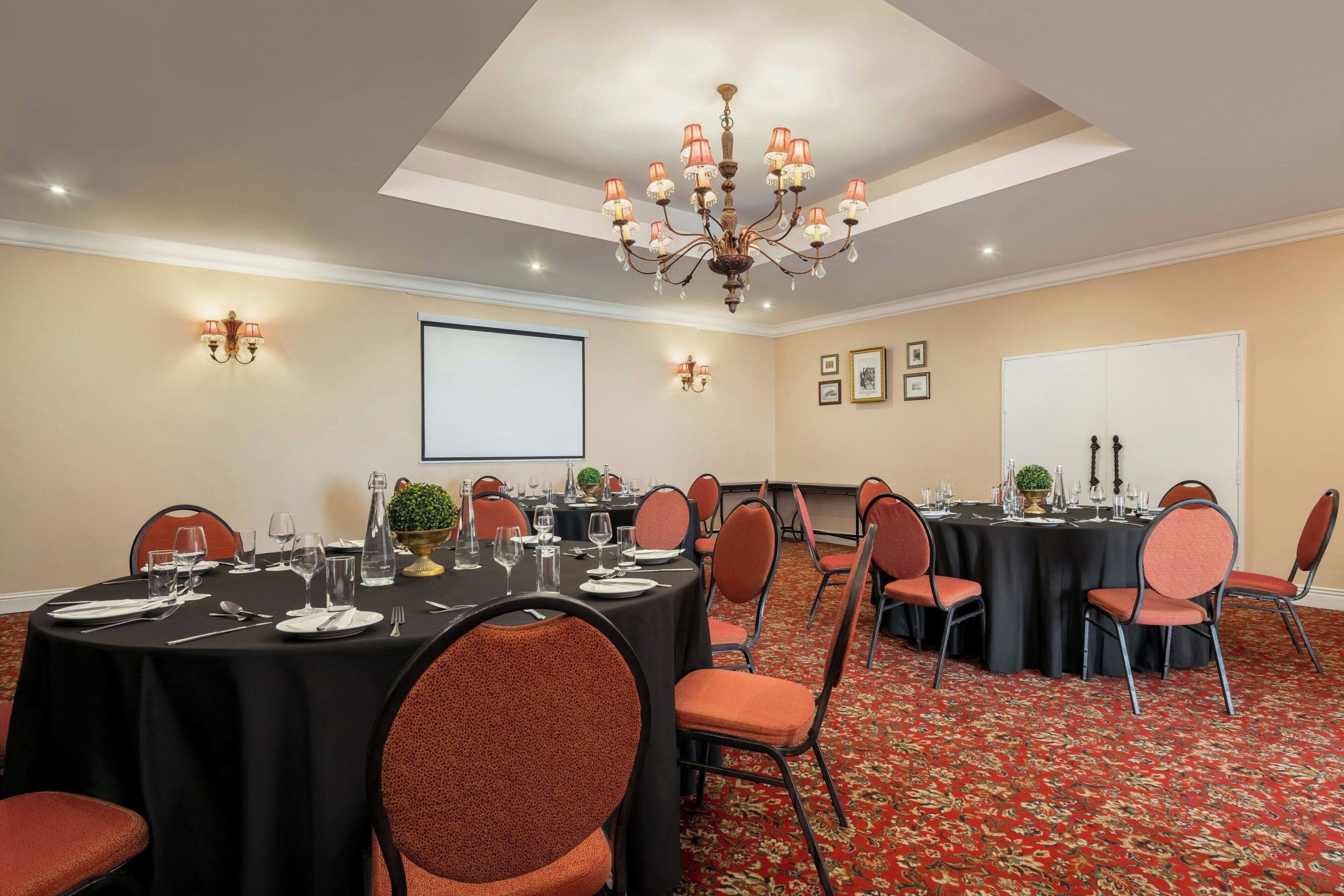A conference room with multiple tables, chairs, a chandelier, and a projector screen.