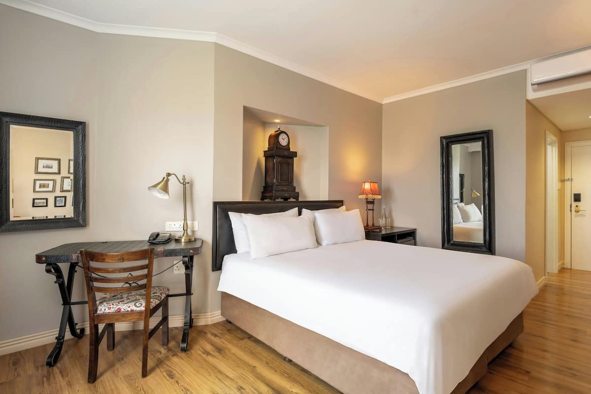 A hotel room with a large bed, a desk with a chair, a clock, a mirror, and a lamp.