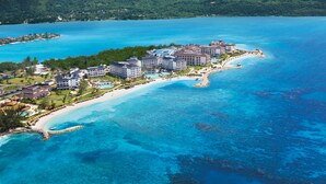 An aerial view of a coastal resort with multiple buildings, a beach, and clear blue waters.