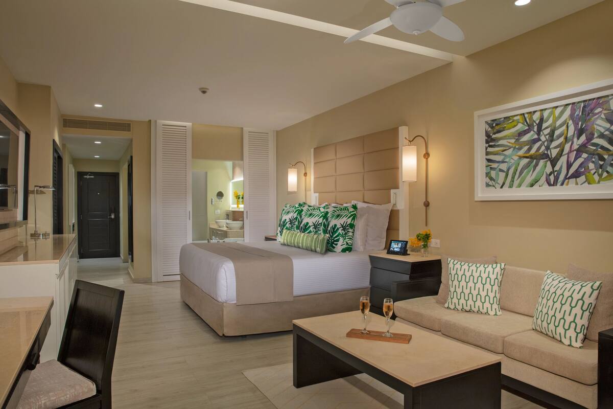 Ein Hotelzimmer mit einem Bett, einem Schlafsofa, einem Couchtisch und einem Schreibtisch. Es gibt einen Deckenventilator und ein Bild an der Wand.