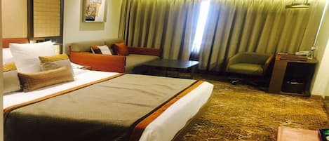 Ein Hotelzimmer mit einem großen Bett, einer Couch, einem kleinen Tisch und einem Stuhl. An der Wand hängt ein Poster und es gibt ein Fenster mit Vorhängen.