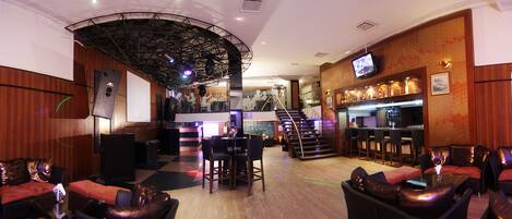 Un área de bar o salón con un escenario, asientos y una escalera que conduce a un nivel superior.