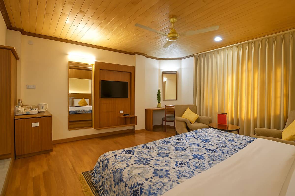 A hotel room with a wooden ceiling, a bed with a blue and white patterned bedspread, a desk with a chair, a television mounted on the wall, and a window with curtains.