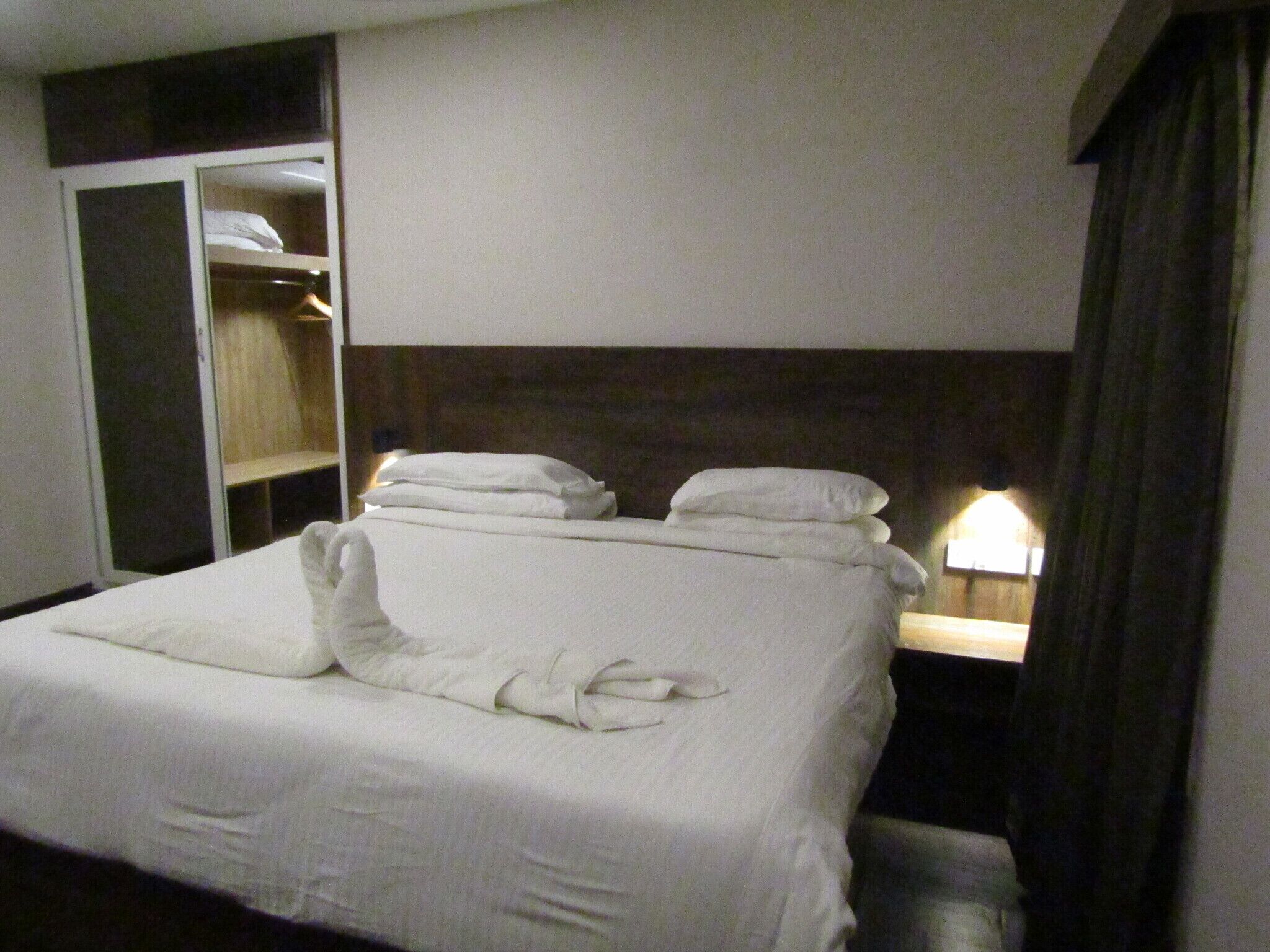 A hotel room with a neatly made bed, a wardrobe, and a bedside table with a lamp.