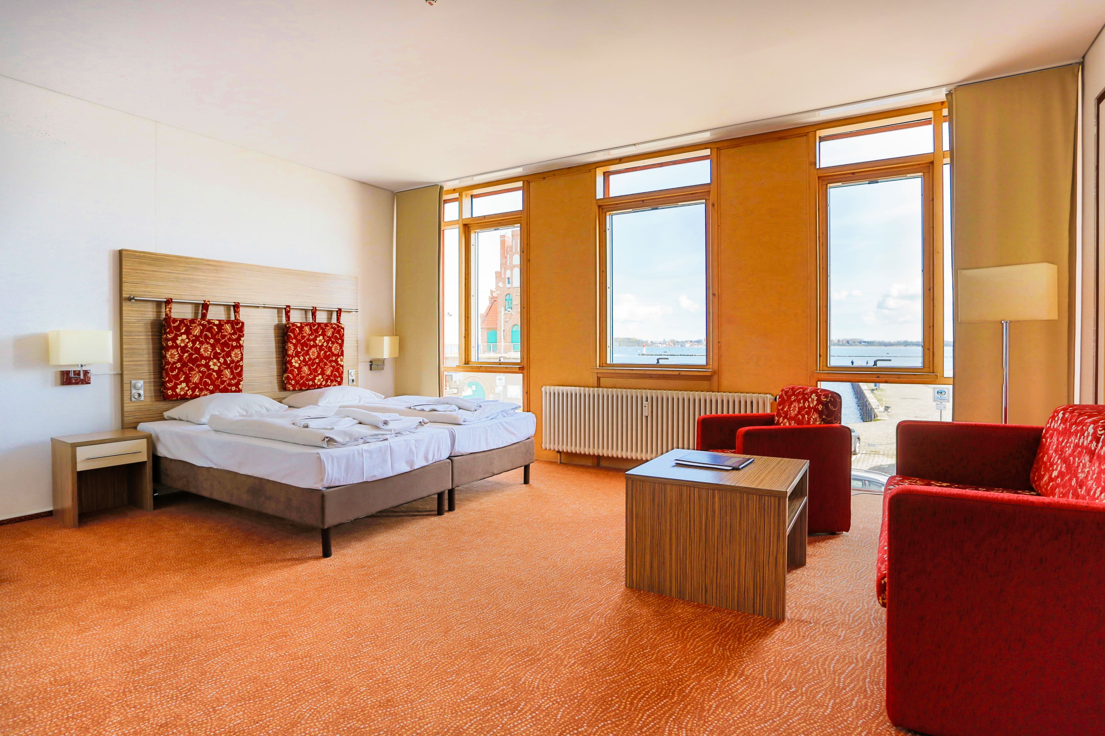 Une chambre d’hôtel avec un lit, un bureau, un fauteuil rouge et de grandes fenêtres donnant sur la ville.
