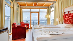 A hotel room with a large bed, a red sofa, a small table, and a view of the city through large windows. - Hotel Hiddenseer (Stralsund)