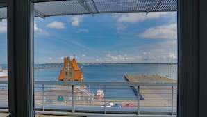 A view from a balcony overlooking a waterfront with a distinctive orange building, a pier, and a boat docked at the pier. - Hotel Hiddenseer (Stralsund)