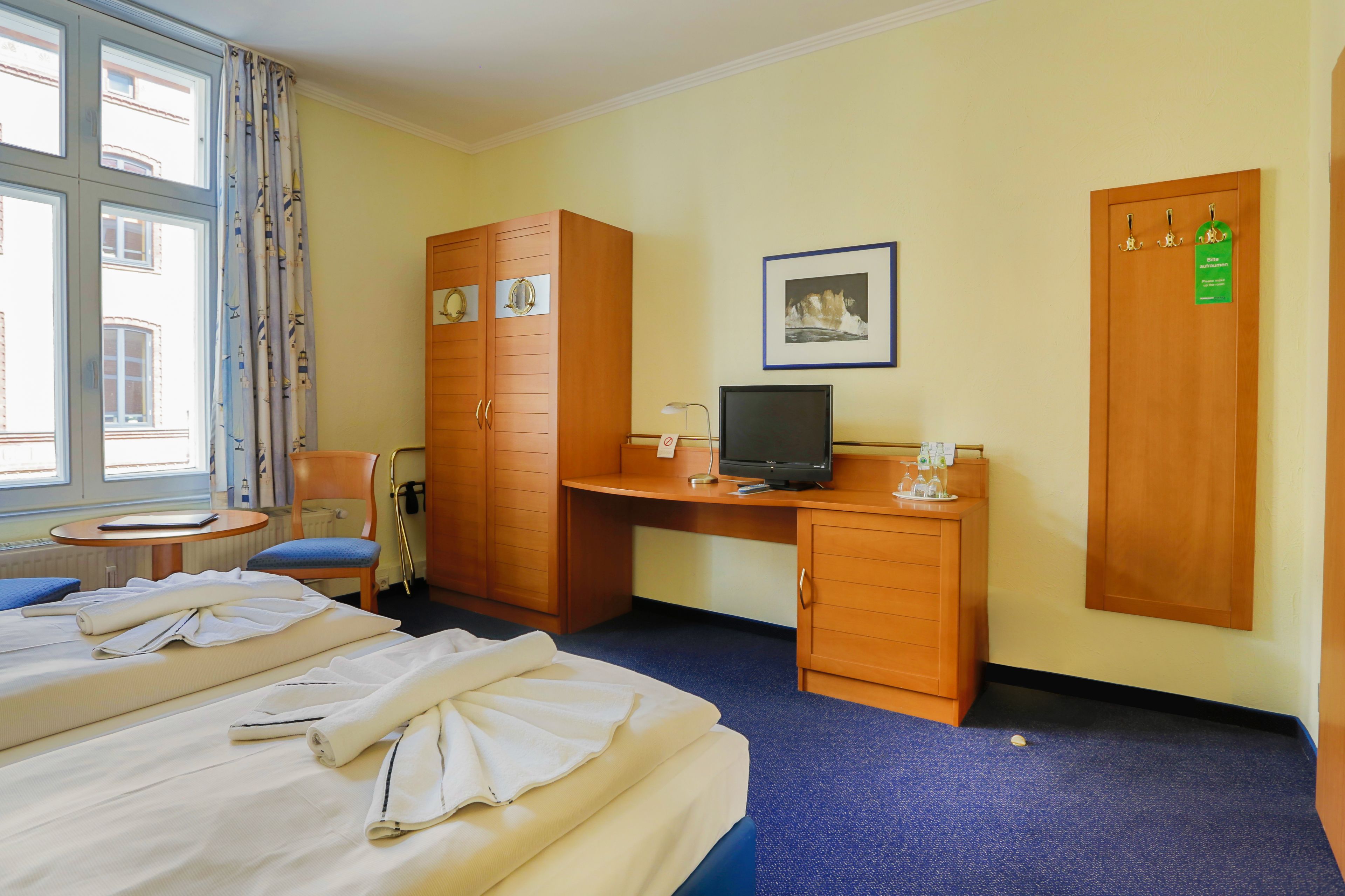 A hotel room with two beds, a desk with a computer, a chair, a wardrobe, and a window with curtains.