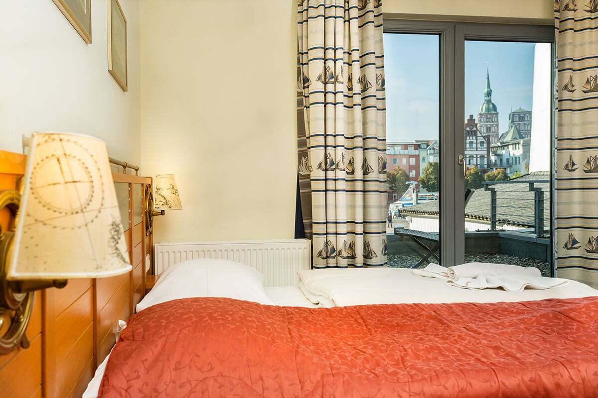 Habitación de hotel con cama, mesita de noche, lámpara y vista a un paisaje urbano a través de una ventana.
