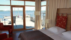 A hotel room with a large window offering a view of the marina and boats. There is a bed with white linens, a red chair, and a small table with a lamp. - Hotel Hiddenseer (Stralsund)