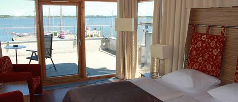 Habitación de hotel con un amplio ventanal que ofrece vistas al puerto deportivo y las embarcaciones. Hay una cama con ropa blanca, una silla roja y una mesita de noche con una lámpara.