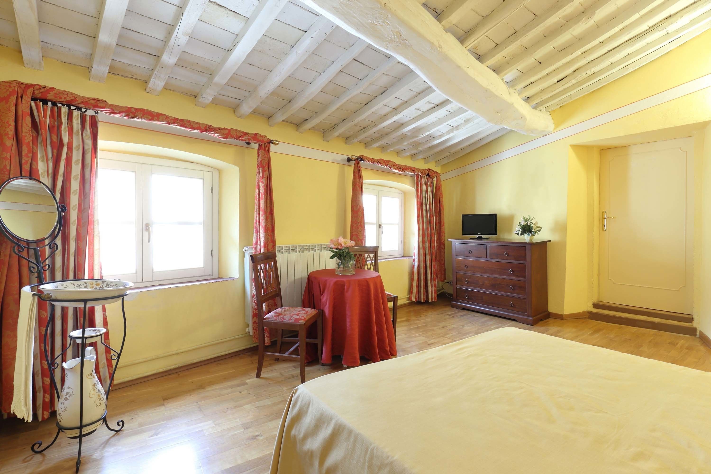 Un dormitorio con cama, cómoda, mesa con un florero, silla y ventana con cortinas.