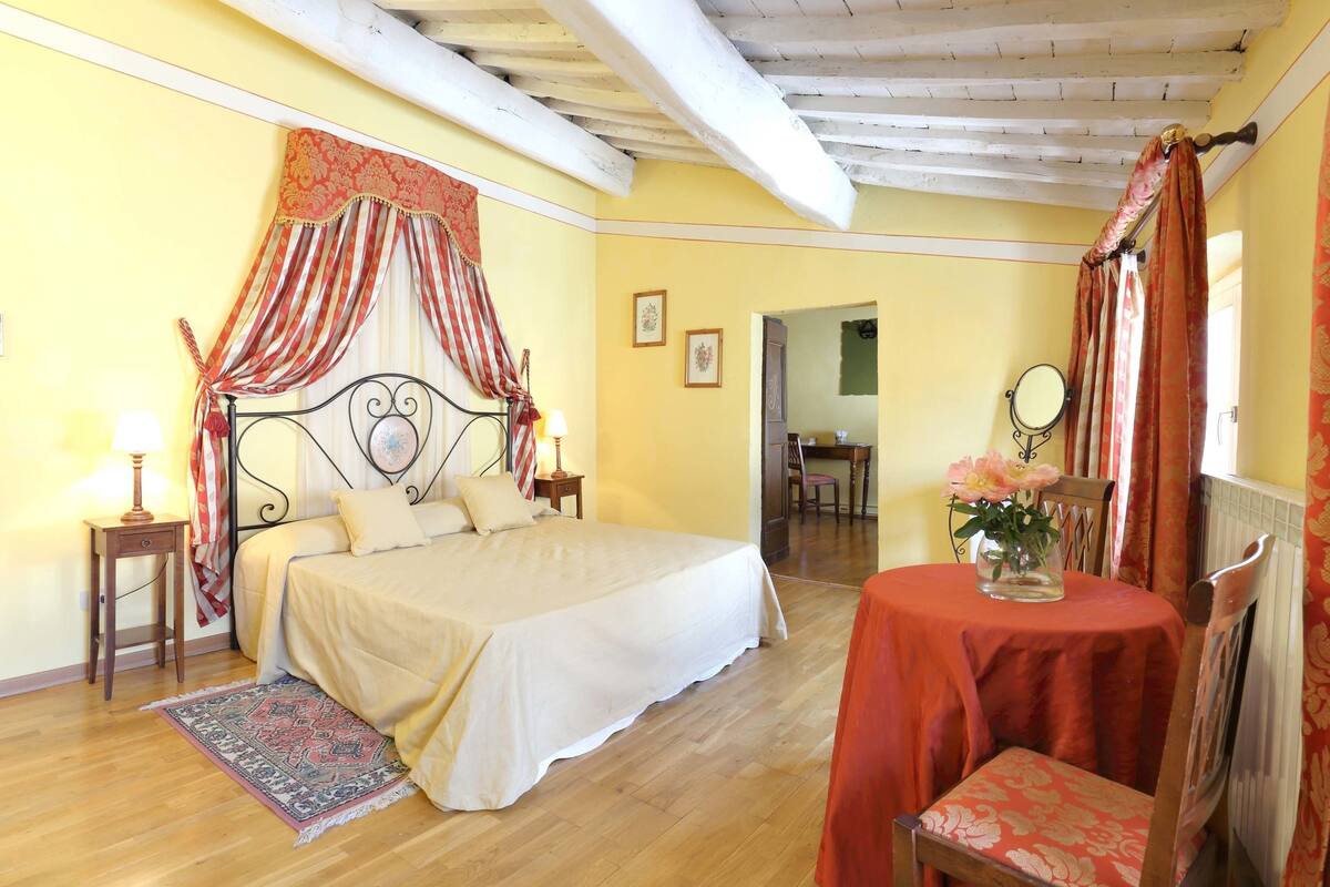 A bedroom with a bed, a bedside table with a lamp, a chair, a table with a vase of flowers, and a mirror on the wall.