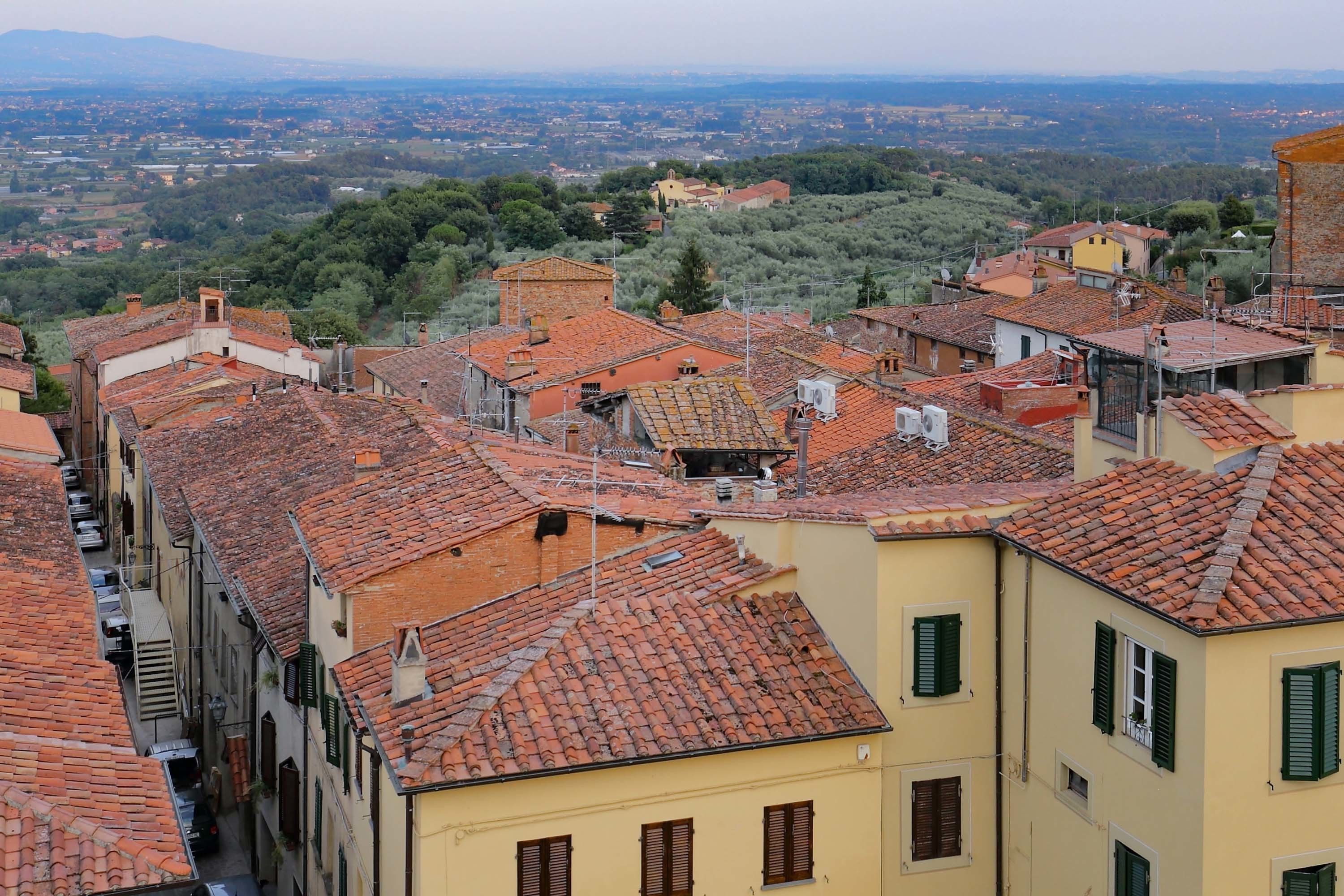 A narrow street with terracotta-tiled buildings, some with green shutters, and a view of a distant town and hills.