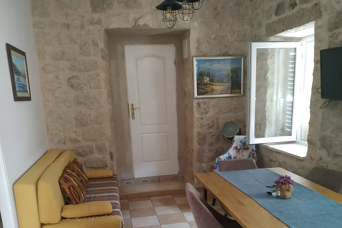 Una habitación con pared de piedra, un sofá amarillo, una mesa de comedor de madera con sillas, una puerta blanca y una ventana con persianas.