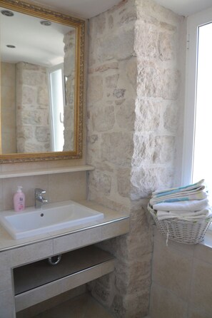 Un baño con pared de piedra, un lavabo blanco, un espejo con marco dorado y una cesta de toallas.