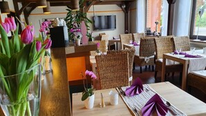 A restaurant with wooden tables and wicker chairs, a bar area with a TV, and a large window with a view. - Beauty & Wohlfühl Hotel (Syke)