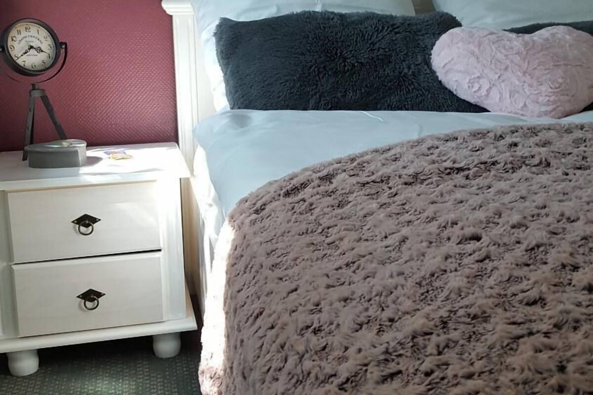 Un dormitorio con cama, almohadas, mesita de noche, reloj y un cuadro floral en la pared.