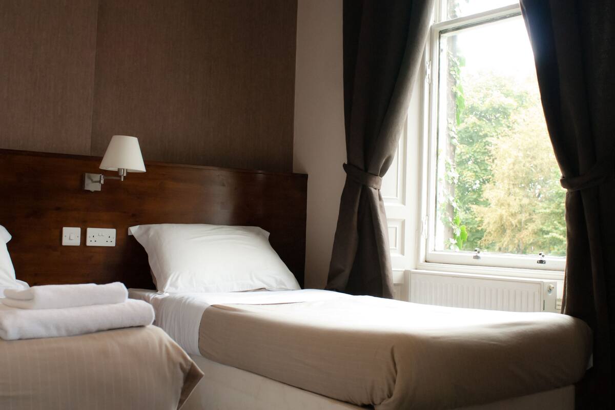 A hotel room with two beds, a wooden headboard, a bedside lamp, and a window with curtains.