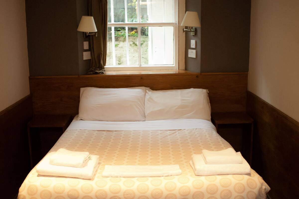 A neatly made bed with a patterned bedspread, two bedside lamps, and a window with a view of greenery outside.