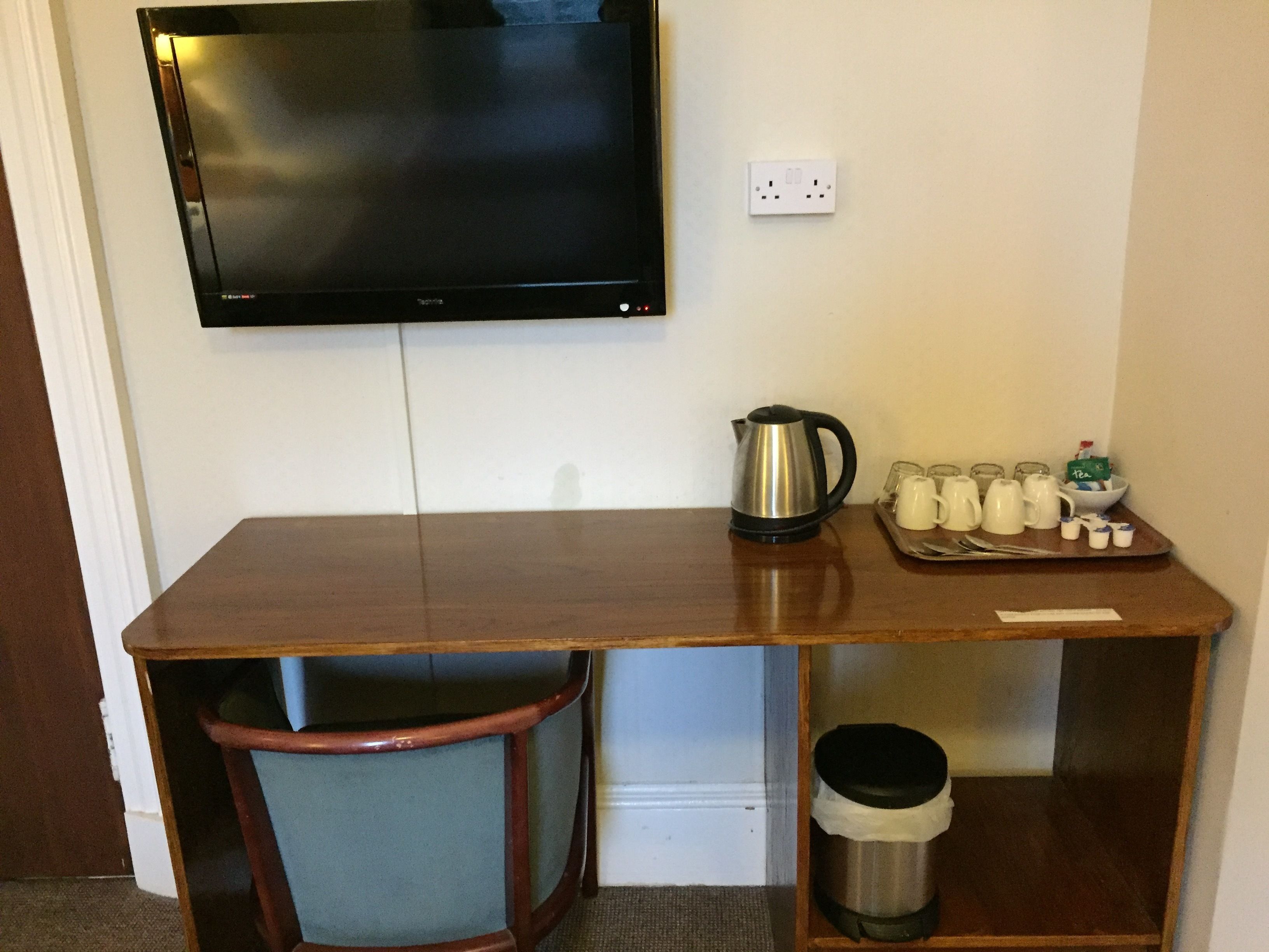A hotel room with a wooden desk, a television mounted on the wall, a kettle, and a tray with cups and a bowl.