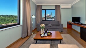 Una sala moderna con un ventanal grande, un sofá gris, una mesa de centro de madera con una bandeja de fruta y un jarrón de flores, y un televisor sobre un mueble de madera.