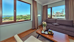 Habitación con un ventanal grande que ofrece vistas a una mezquita y vegetación. Hay un sofá, una mesa de centro con una bandeja de fruta y un jarrón con flores.