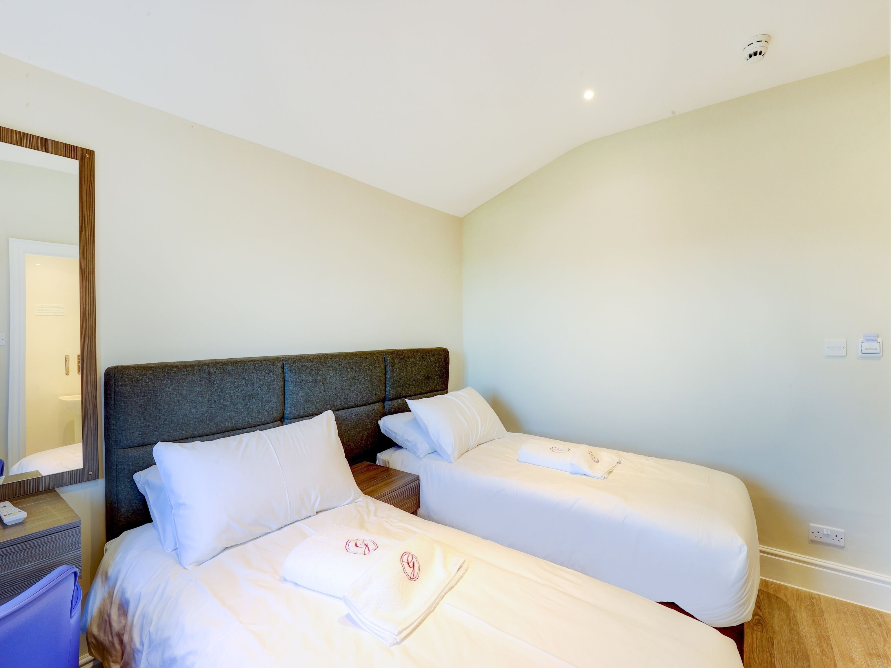 A hotel room with two single beds, a bedside table, a mirror, and a light switch on the wall.