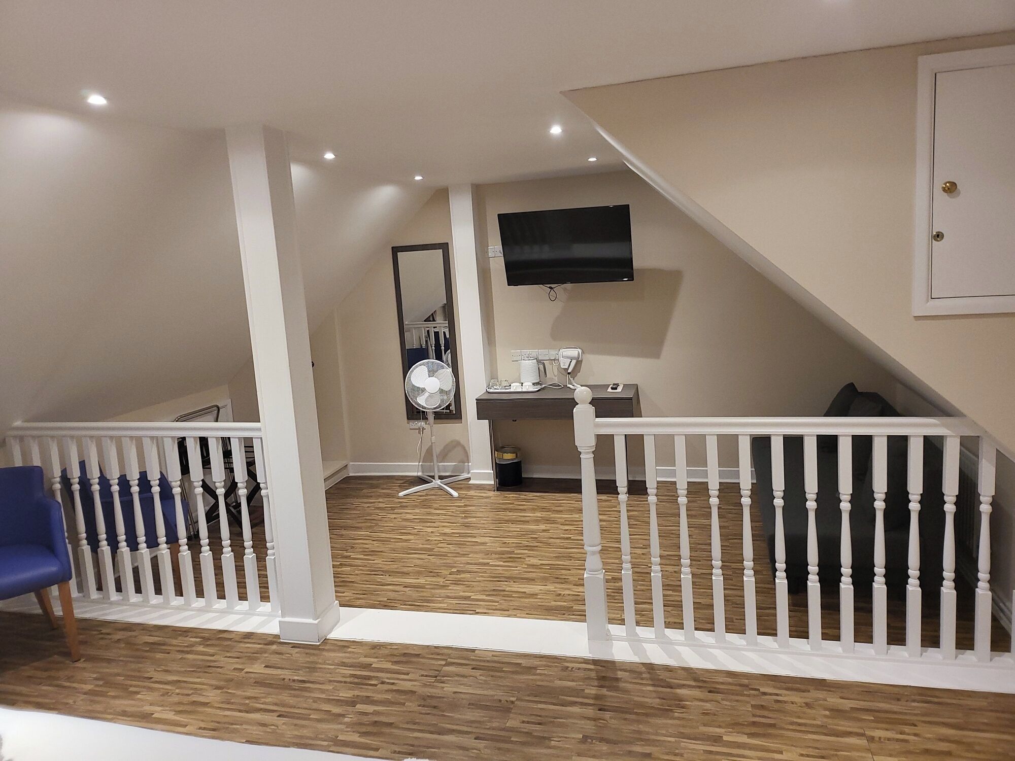 A room with a wooden floor, white railings, a blue chair, a desk with a computer, and a TV mounted on the wall.