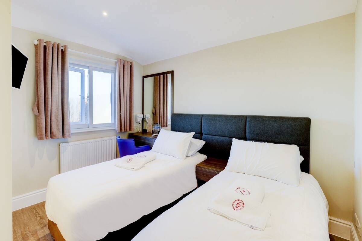 Ett hotellrum med två sängar, ett fönster med gardiner, en spegel, ett skrivbord med en telefon och vattenflaskor samt en blå stol.